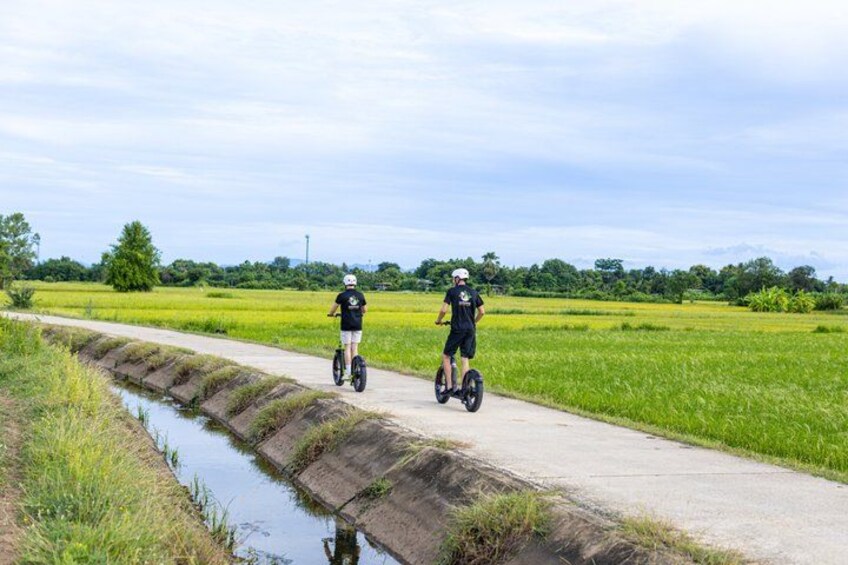 Two people riding Segways on a paved path alongside a water channel in a rural landscape. 1