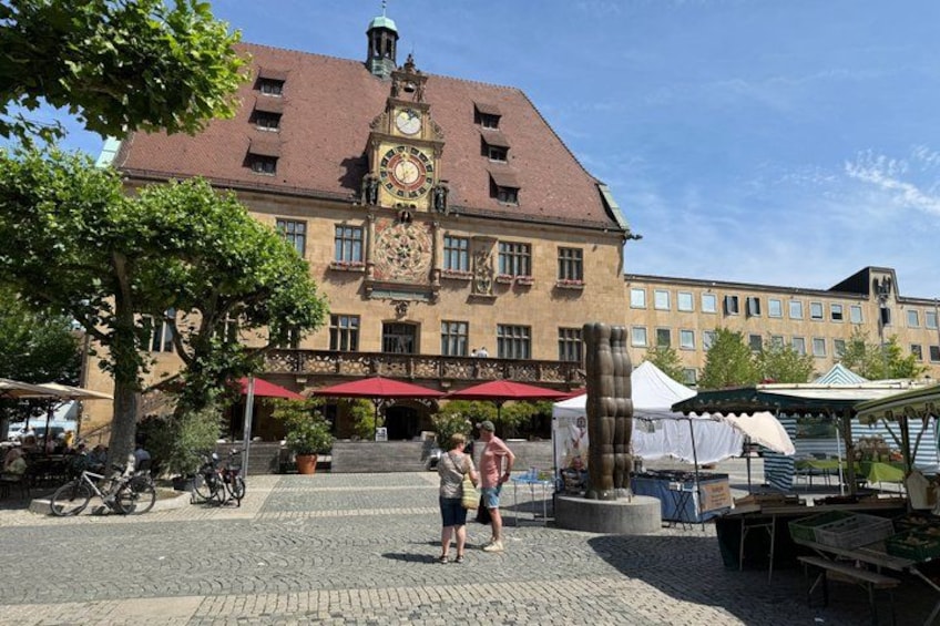A historic building with a clock tower, cobblestone plaza, and outdoor market stalls. 2