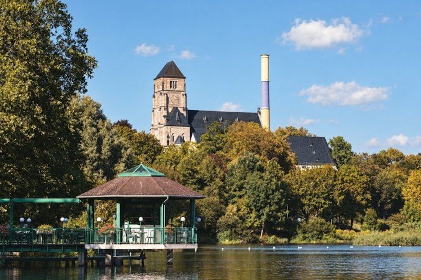 A historic building with a tower and chimney, a gazebo by a lake, and trees in the background. 5
