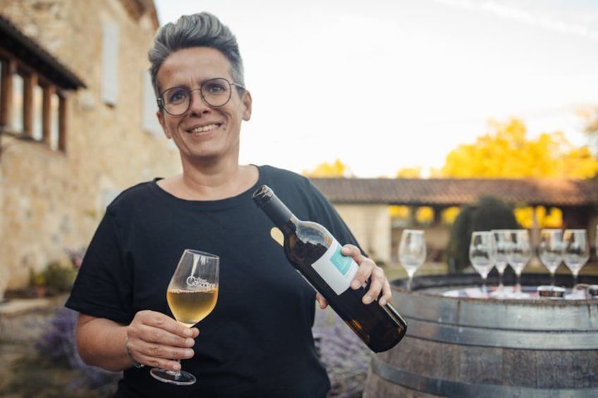 A person holding a wine bottle and a glass of wine, with a barrel and a stone building in the background. 3