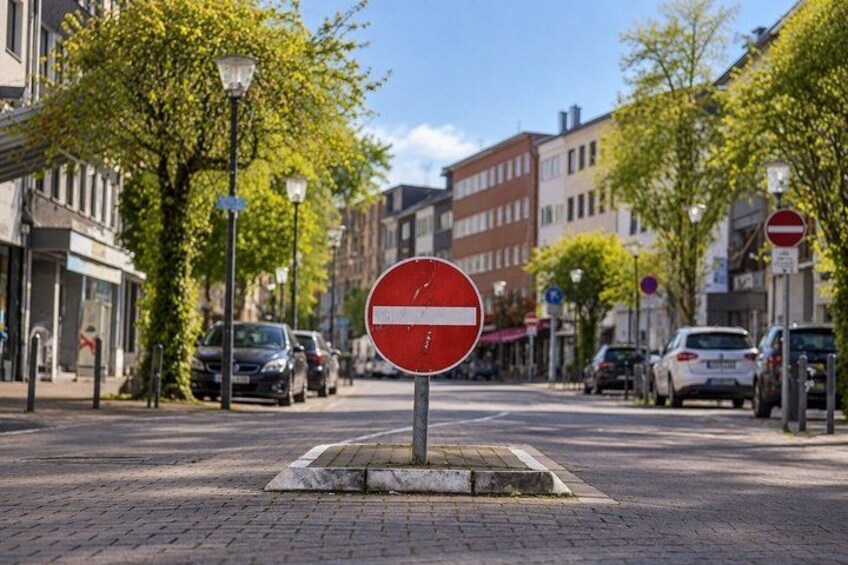 A no entry road sign on a paved street with cars parked on the side, trees, and buildings in the background. 3