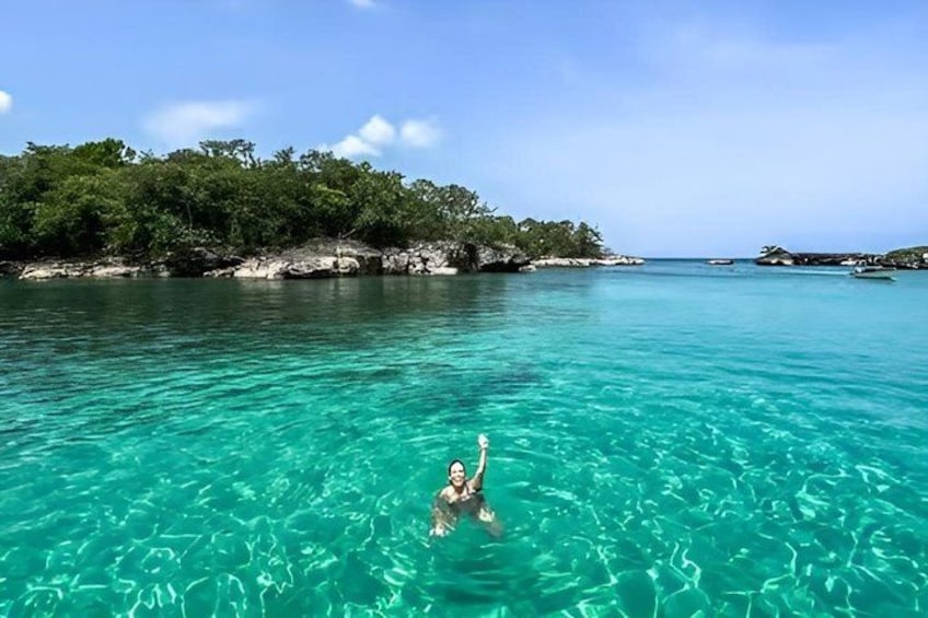 A person swimming in clear turquoise water with a rocky coastline and trees in the background. 1
