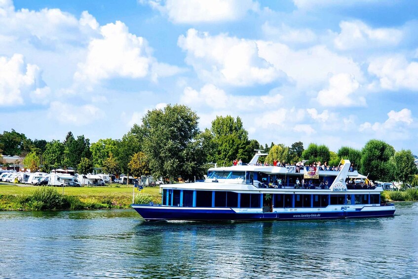 A blue and white river cruise boat with passengers on deck, surrounded by greenery and parked RVs on the shore. 4