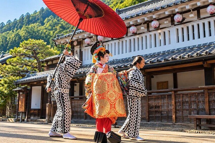 A person in traditional Japanese attire holding a red fan, standing in front of a traditional Japanese building with a tiled roof and wooden structures. 1