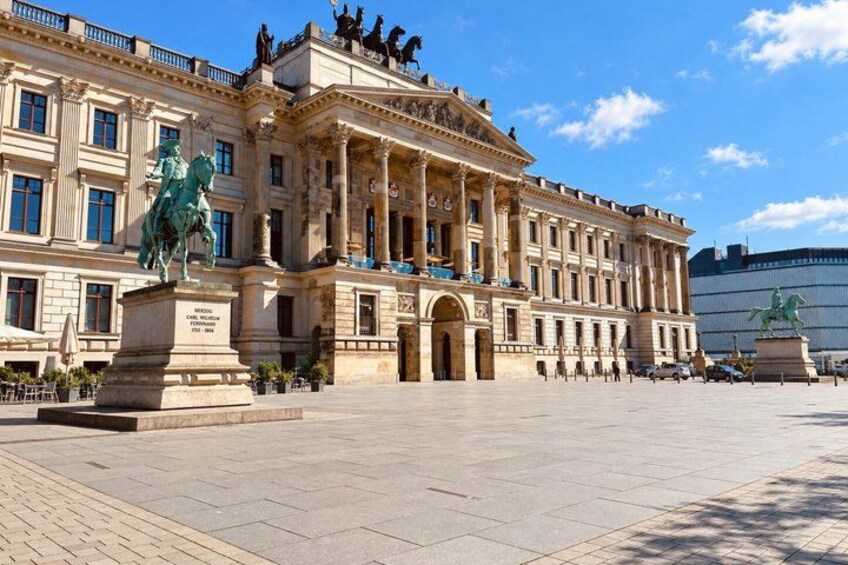 A large classical building with columns and statues, a wide open plaza, and a clear blue sky. 4