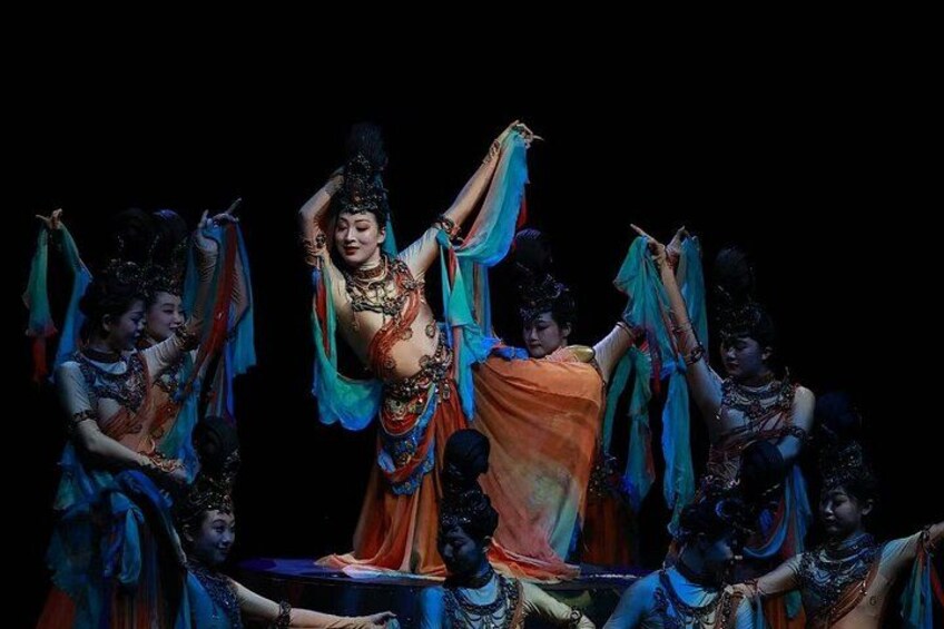 A group of performers in traditional costumes, with one central figure in a flowing orange garment and others in blue and orange attire, performing a dance. 4