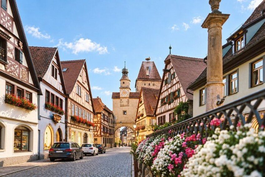 A cobblestone street lined with traditional half-timbered houses, a church tower, and a flower-adorned fence. 2