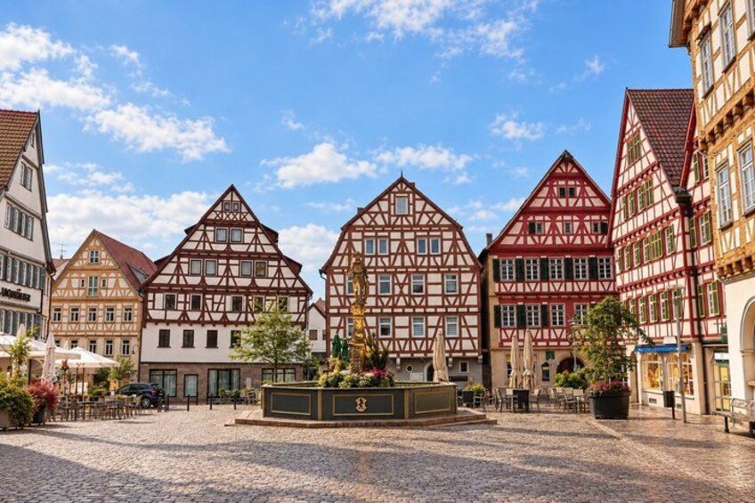 A cobblestone square with traditional half-timbered houses, a fountain, and outdoor seating. 4