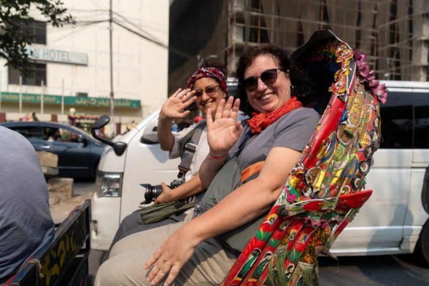 A person in a colorful, patterned outfit sitting in a vehicle, with a building and a car in the background. 1
