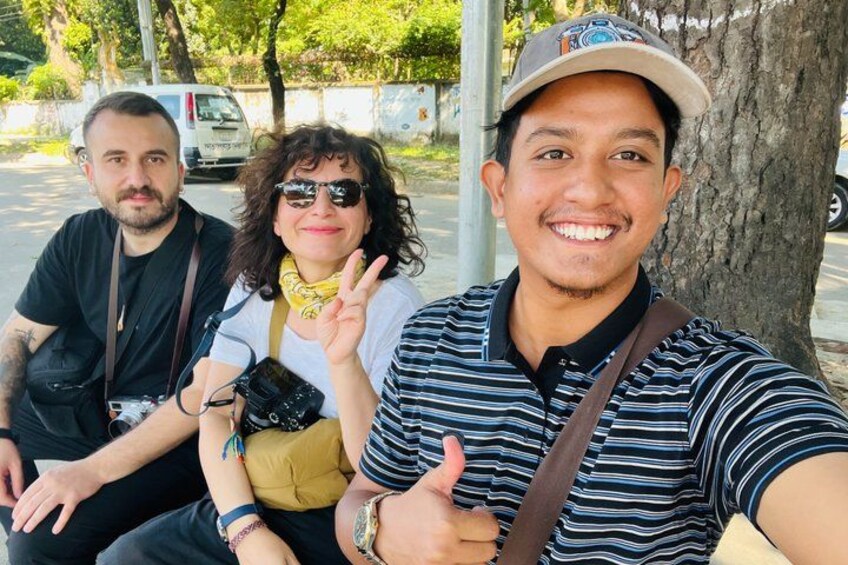 Three people sitting on a bench with a tree and a parked car in the background. 2