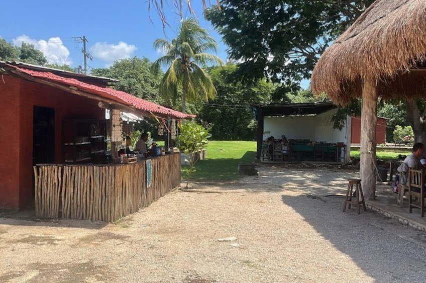 A rustic outdoor dining area with a thatched-roof structure, a wooden fence, and a gravel ground. 3