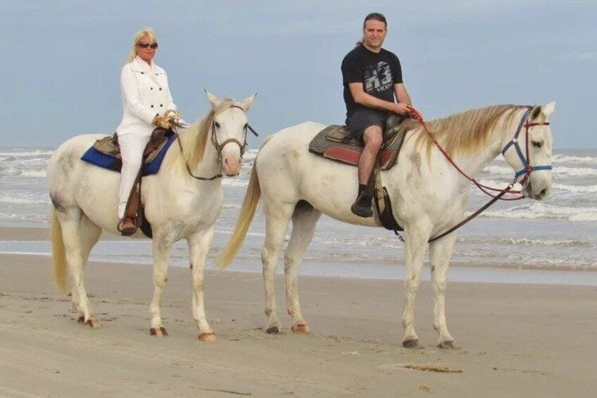 Two people riding horses on a beach with the ocean in the background. 2