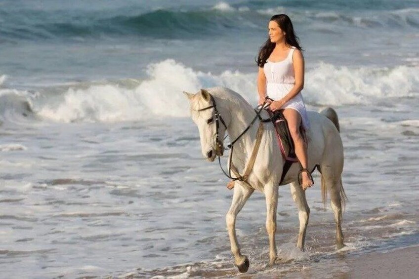 A person riding a white horse along a beach with waves in the background. 1
