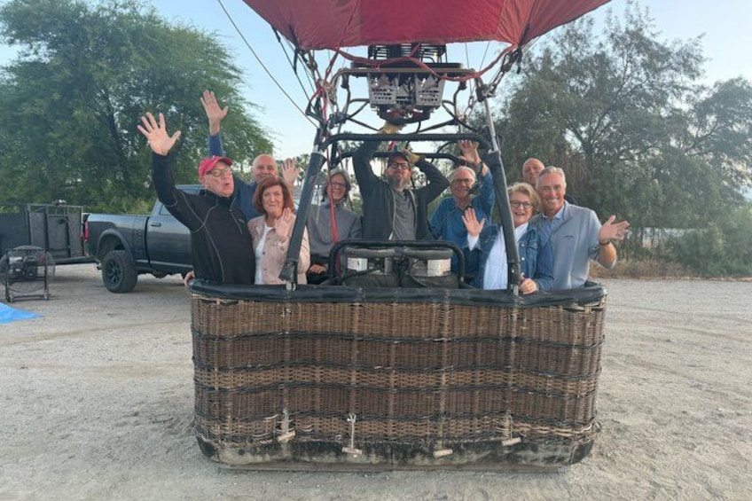 A group of people in a wicker hot air balloon with a red canopy, parked on a gravel surface. 5