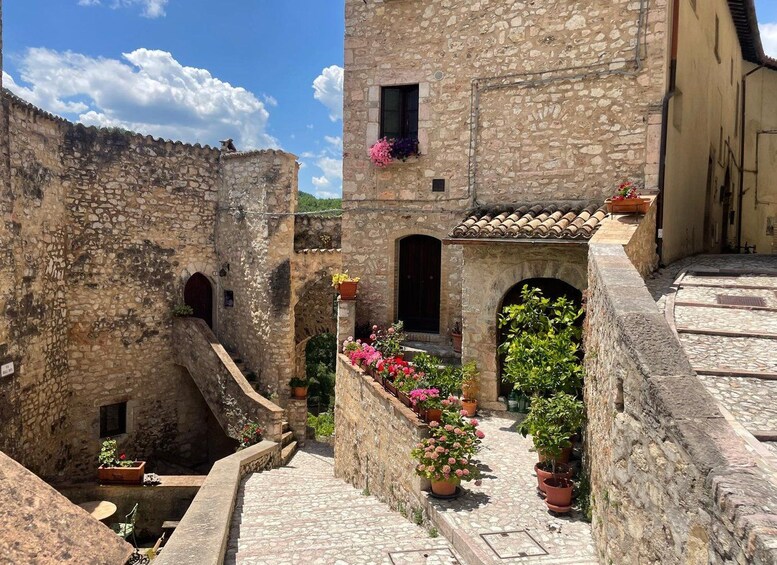 A stone building with a tiled roof, a staircase leading to an upper level, and potted plants with flowers. 2