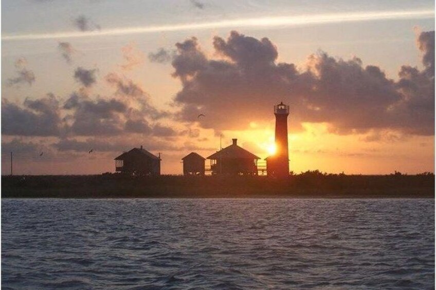 A lighthouse, two small buildings, and a sunset over water. 1