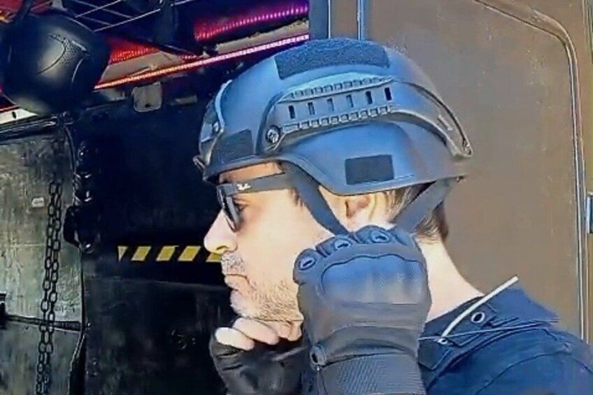 A person wearing a tactical helmet and gloves, standing in front of a vehicle with a chain and a red light. 5