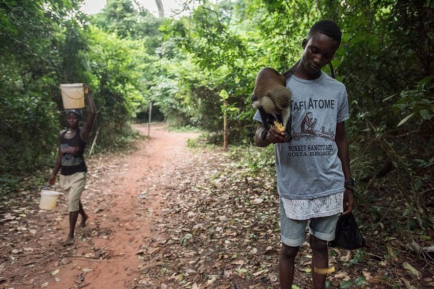Two individuals walking on a dirt path in a forested area, one carrying a bucket and the other holding a baby. 1