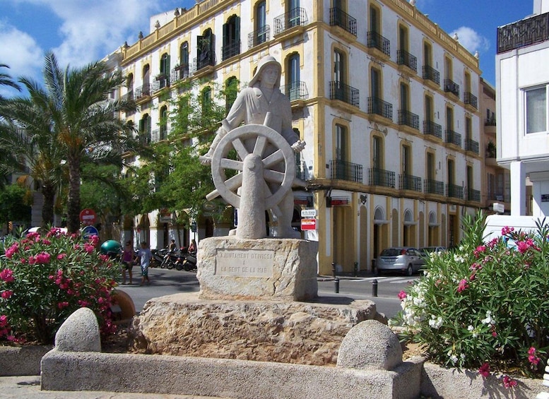 A statue of a woman with a ship's wheel, surrounded by flowers and palm trees, in front of a yellow building with balconies. 3
