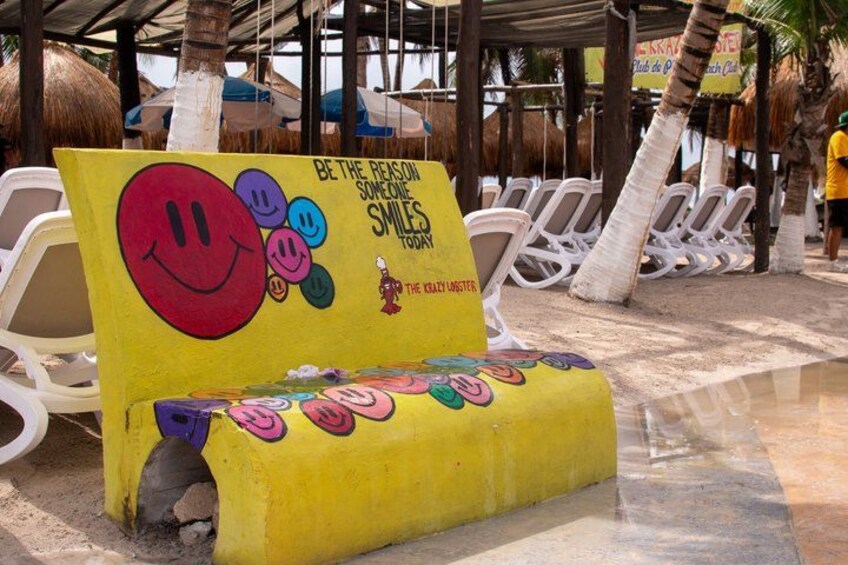 A yellow bench with colorful smiley faces and text, located in a beachside area with white lounge chairs and palm trees. 4