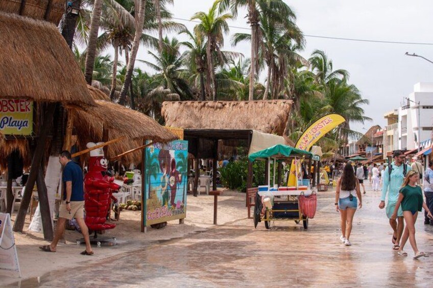 A tropical street with thatched-roof stalls, palm trees, and people walking. 2
