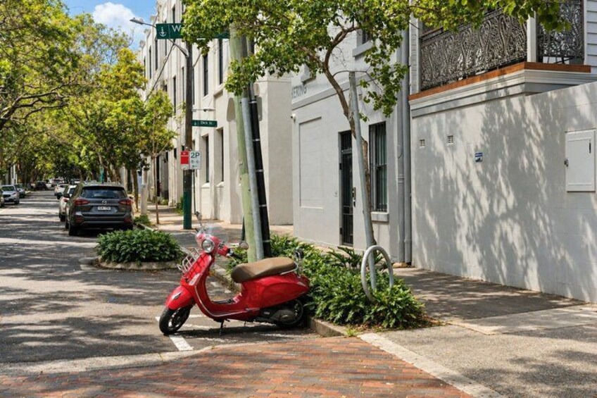 A red scooter parked on a brick sidewalk, a tree, and a row of white buildings with a street in the background. 3