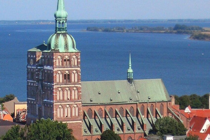 A historic church with a tall tower and a smaller building with a green roof, set against a backdrop of a large body of water and distant land. 3