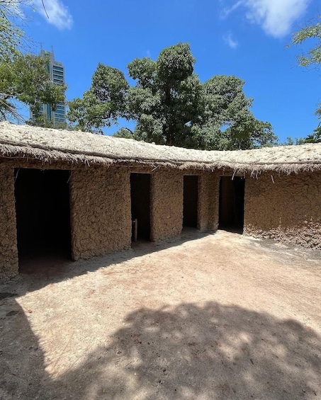 A traditional thatched-roof structure with multiple openings, surrounded by a dirt ground and trees. 2