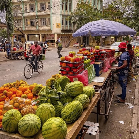 A street market with fresh produce like watermelons and oranges, a man on a bicycle, and a vendor under a blue umbrella. 3