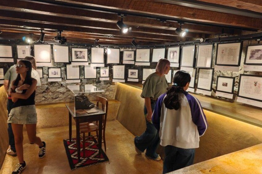 An indoor exhibit with framed pictures on the walls, a wooden bench, and a table with a chair. 5