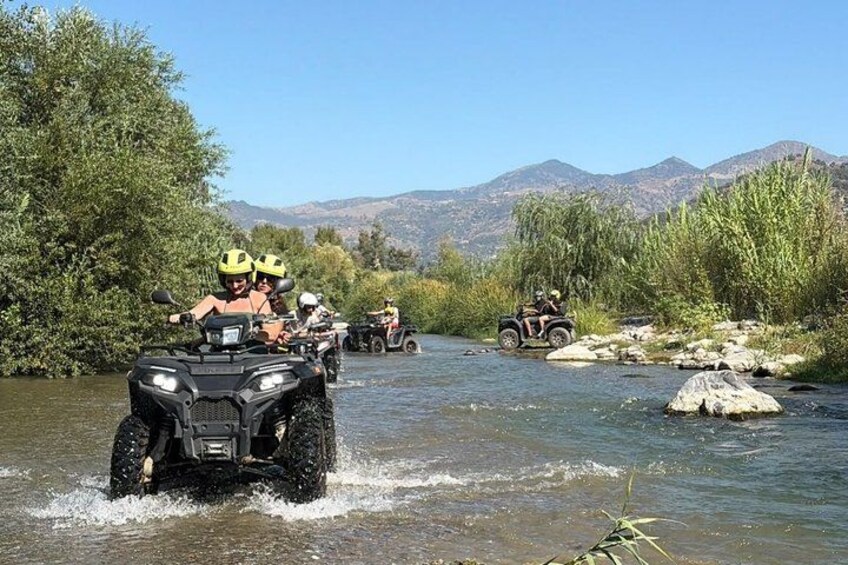 People riding ATVs on a river surrounded by trees and mountains. 3