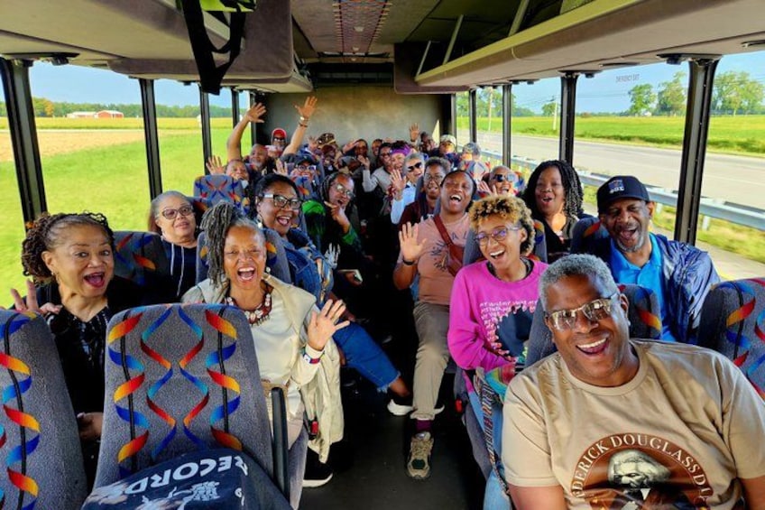 A group of people on a bus, some raising their hands, with a rural landscape visible through the windows. 1
