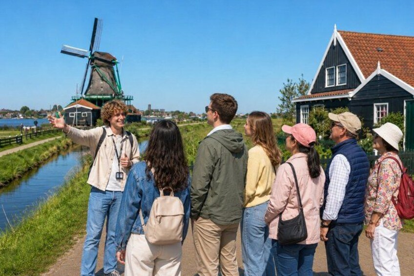 A group of people standing on a path near a windmill and a house by a canal. 1