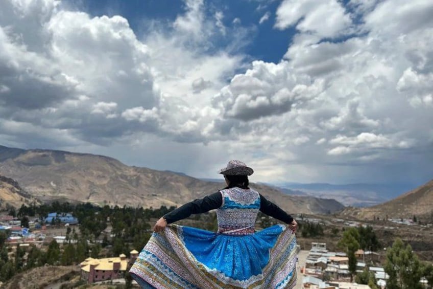 A person in traditional attire standing with arms outstretched, overlooking a mountainous landscape with scattered buildings. 1