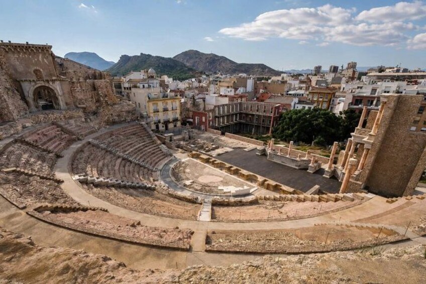 An ancient amphitheater with ruins, surrounded by modern buildings and mountains in the background. 1