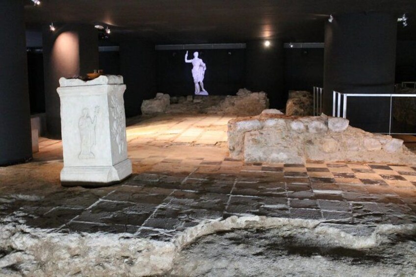 An archaeological site with stone slabs, a pedestal with carvings, and a projected image of a figure. 4