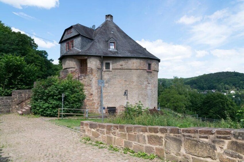A stone tower house with a steep roof, surrounded by greenery and a stone wall. 3