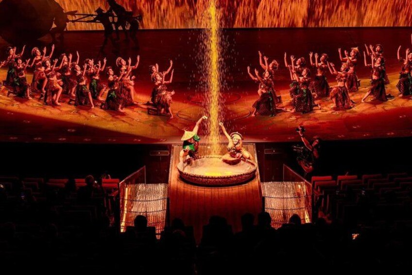 A stage performance with dancers in traditional costumes, a central fountain, and an audience seated in rows. 1