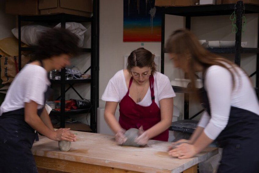 Three people working at a wooden table with flour, in a room with shelves and a colorful wall. 3