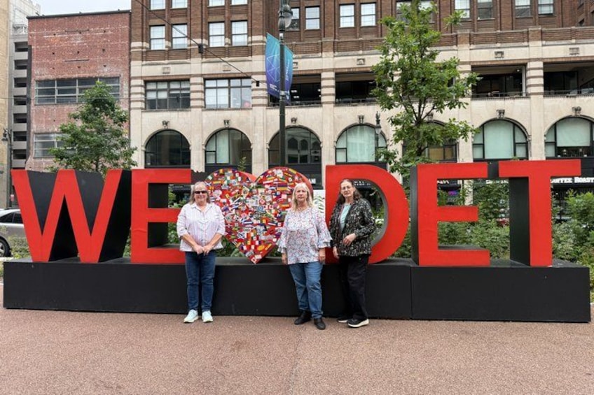 Three people standing in front of a large sign with the letters 'WEDE' and a heart-shaped mosaic. 2