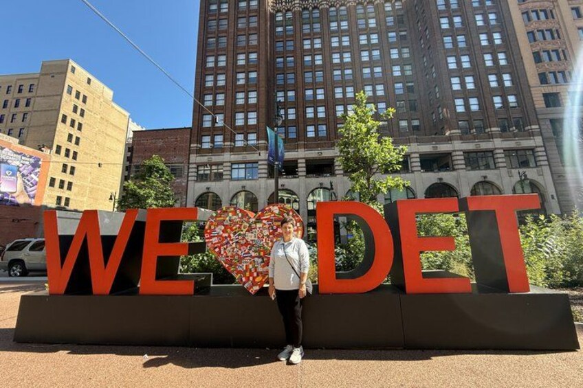 A person standing in front of a large sign with the words 'WE ♥ DET' and a heart-shaped design. 3