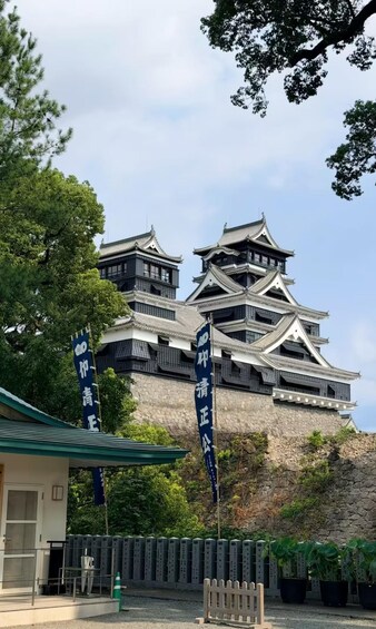 A traditional Japanese castle with multiple tiers and a stone base, surrounded by greenery and featuring two prominent banners with Japanese characters. 2