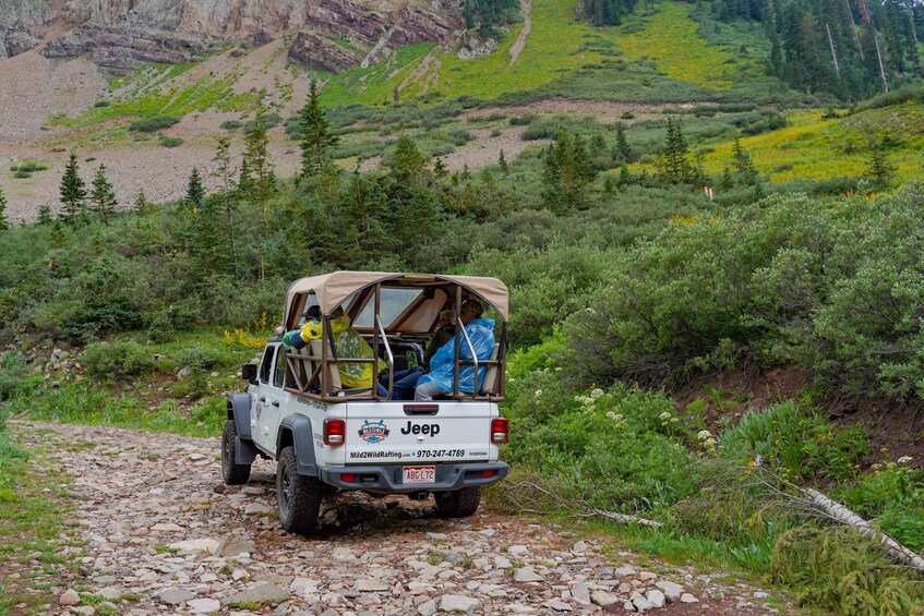 A Jeep with a canvas cover and a person inside, parked on a rocky trail surrounded by greenery and mountains. 2