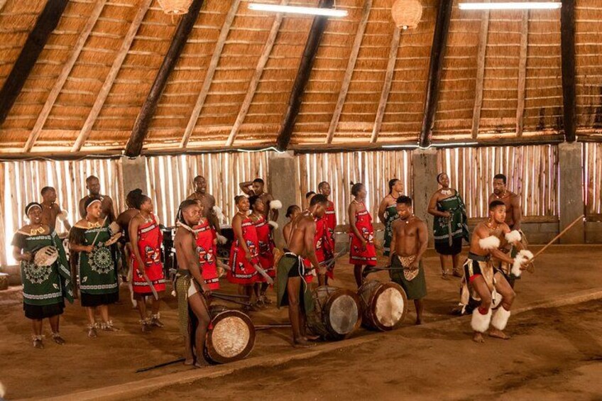 A group of people in traditional attire, some playing drums, in an indoor setting with a thatched roof. 1