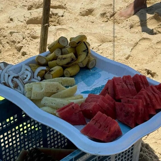 Une scène de plage avec un panier en plastique contenant des tranches de pastèque, des rondelles de banane et des oignons frits. 4