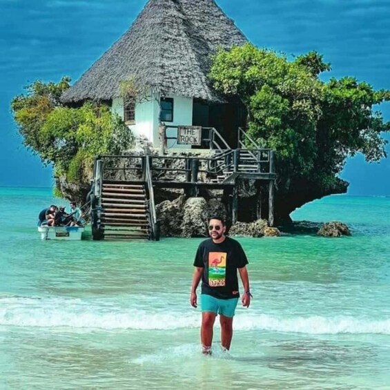 Une personne se tient dans une eau peu profonde, devant une maison au toit de chaume, sur une île rocheuse aux eaux d’un bleu limpide. 5