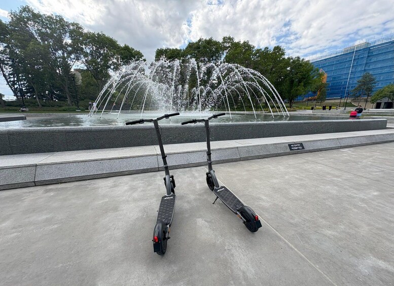 Two electric scooters parked in front of a fountain with water jets. 1
