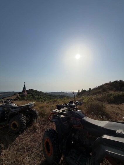 A sunny landscape with two ATVs parked on a grassy hill, a flag, and a distant hill with a structure. 2