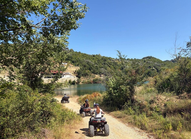 A dirt path with people riding ATVs, a body of water, and a hillside with vegetation. 1