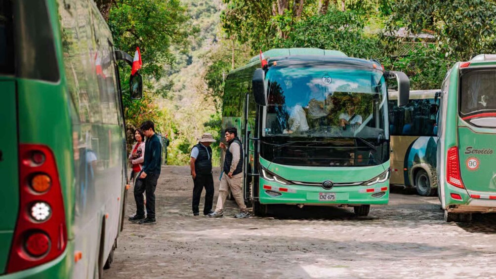 Un groupe de personnes se tient à côté d’un autobus vert portant le numéro 26, qui est garé sur une route de terre, avec d’autres autobus en arrière-plan. 4